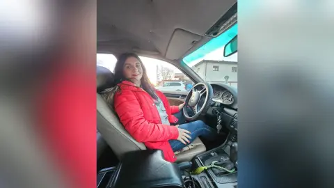 Smiling woman in a red puffer jacket sitting in a car driver's seat on a bright day.