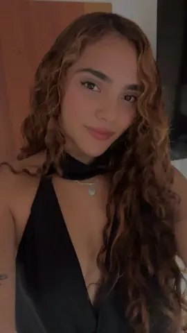 Young woman with long wavy hair wearing a black halter top and silver necklace in a warm indoor setting.