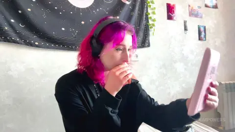 Young woman with pink hair wearing headphones eating snacks while watching a video on a tablet in a cozy room.