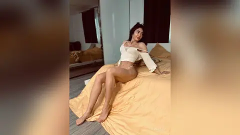 Glamorous woman in white off-the-shoulder crop top and lingerie sitting on a bed in a bright bedroom.