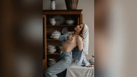 Redheaded woman in a blue striped shirt and denim jeans sitting in a rustic kitchen setting.