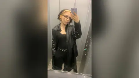 Trendy woman in black leather jacket and glasses taking a mirror selfie in an elevator.