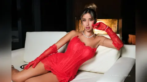 Glamorous woman in a red lace mini dress and matching gloves relaxing on a white sofa in a moody setting.