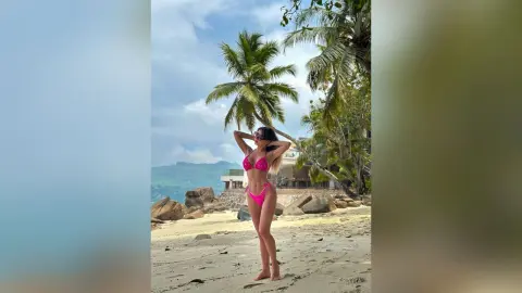 Katia poses in a pink bikini on a tropical beach with palm trees and sand behind her.