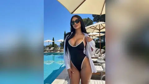 Glamorous woman in a black one-piece swimsuit and white cover-up relaxing by a sunny luxury resort pool.