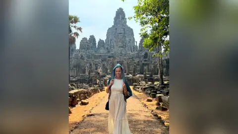 AnaMathers stands on a stone path in front of an ancient stone temple wearing a white dress and a blue headscarf.
