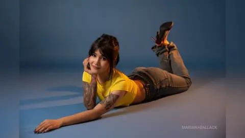 Smiling woman in a yellow t-shirt and jeans lying on a blue studio background in a playful mood.