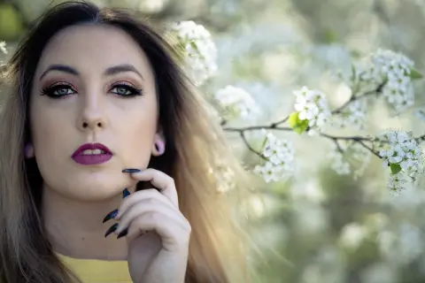 Young woman with glamorous makeup and long hair posing amidst white spring blossoms in a dreamy garden setting.