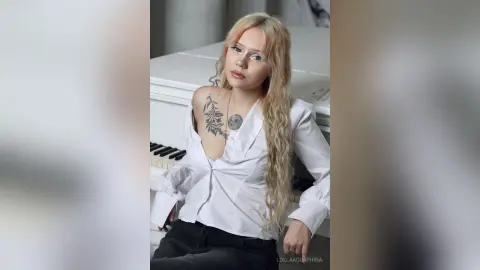 Blonde woman with tattoos in a white button-down shirt sitting by a white piano in a bright, minimalist setting.