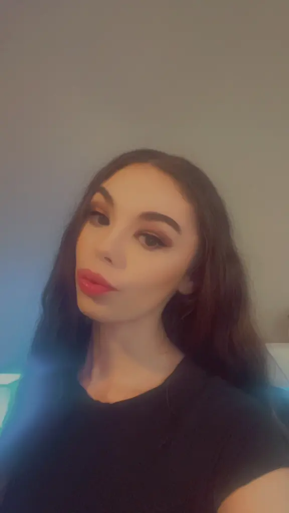 SaraBrynleeXOXO — Young woman with long dark hair and red lipstick wearing a black t-shirt in soft indoor lighting.