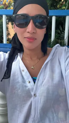 Smiling woman in a white linen shirt and black hijab wearing sunglasses on a sunny outdoor balcony.