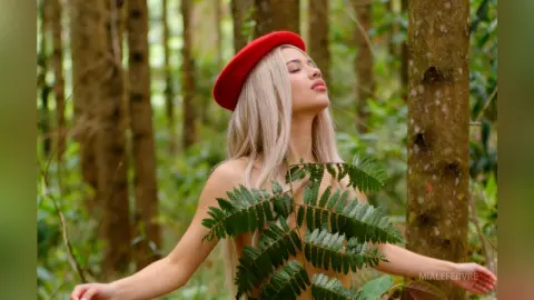 Blonde woman in a red beret posing peacefully amidst lush green forest trees.