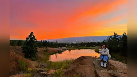 Smiling woman in a white sweater sitting on a rock by a lake under a vibrant orange sunset sky.