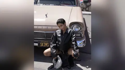 Stylish woman in a black leather jacket sitting by a vintage beige car in bright sunlight.