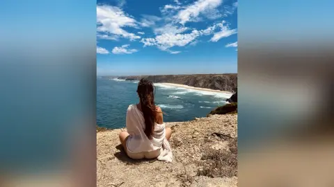 Woman in a white dress sitting on a coastal cliff overlooking the blue ocean under a sunny sky.