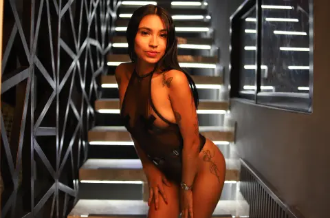 Tattooed woman in black sheer bodysuit posing on modern illuminated stairs in a moody, dark interior.