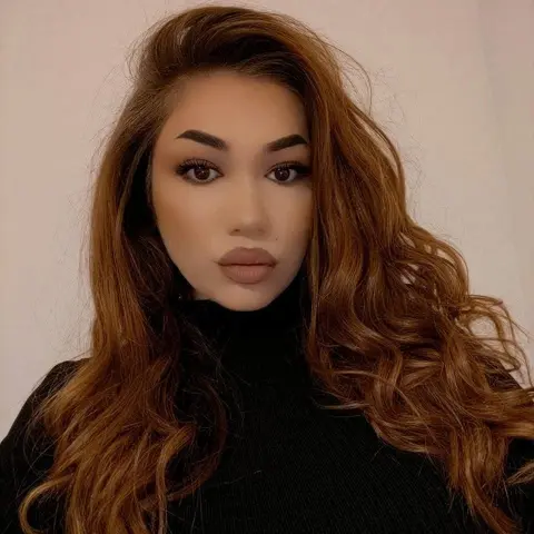 Brunette woman in a black turtleneck sweater posing with long wavy hair against a neutral background.
