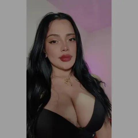 Young woman with long dark hair wearing a black low-cut top and gold necklace in soft indoor lighting.