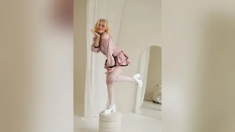 LynGoodhart poses playfully in a pink ruffled dress and white high heels while standing on a white pedestal.