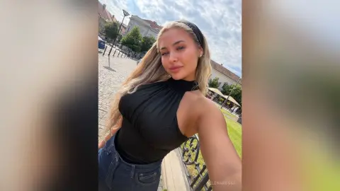 SelenaRusse poses outdoors in a black halter top and blue jeans, looking at the camera with her long blonde hair flowing down.