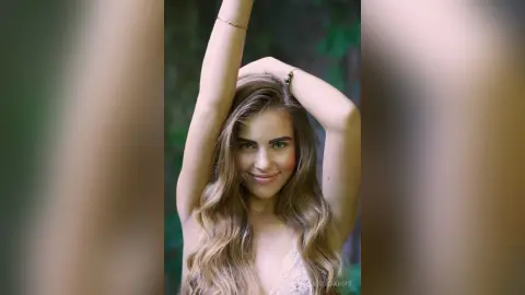 Smiling young woman with long wavy hair in a white lace dress posing in a lush green forest setting.