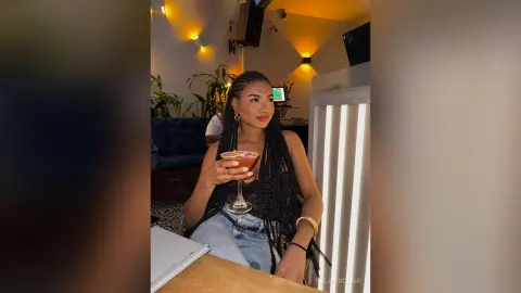 Stylish woman with long braids enjoying a cocktail in a warm, dimly lit lounge setting.