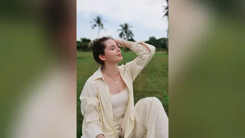 LiaMotzer smiles with her eyes closed, wearing a white tank top and open button-down shirt in a grassy field.