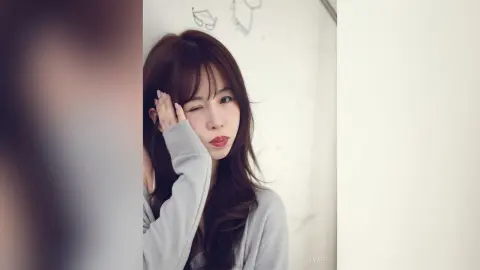 Winking young woman in a grey long-sleeve sweater posing playfully against a white background.