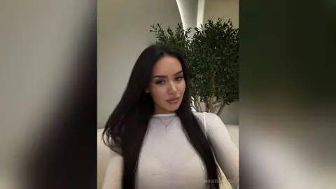 Young woman with long dark hair wearing a white long-sleeve top in a bright, minimalist indoor setting.