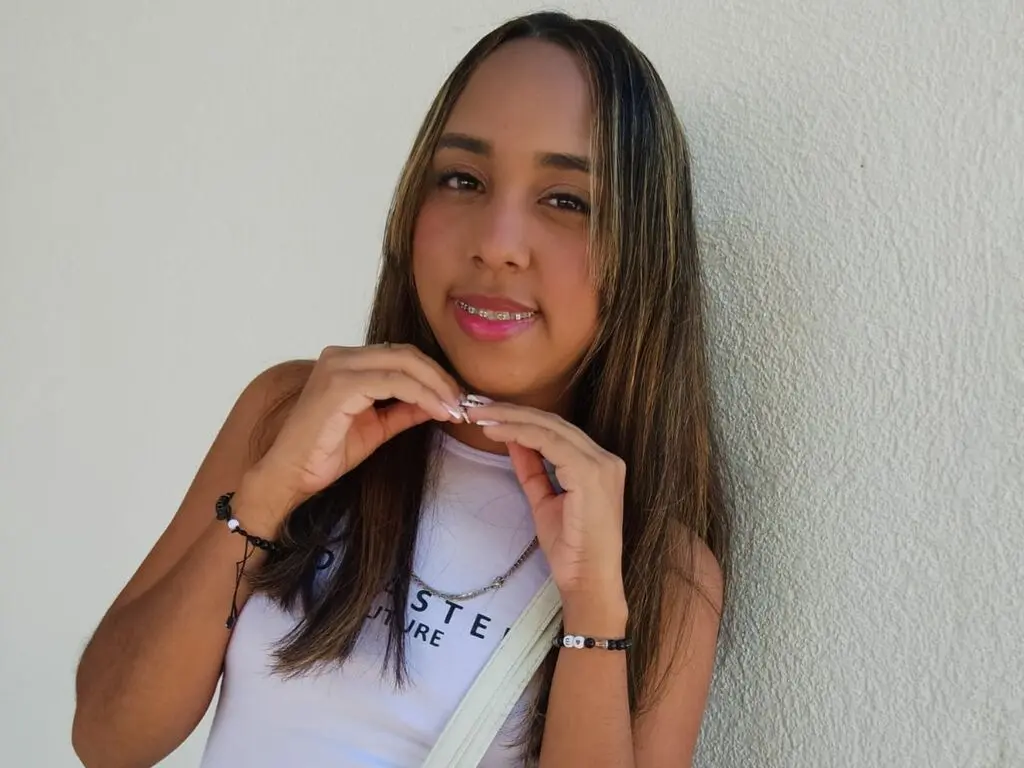 ArianaIvers poses with her hands near her neck, wearing a white tank top and long straight hair against a plain white wall.