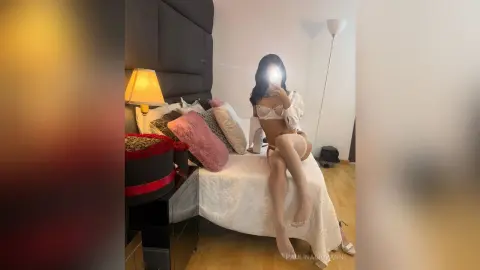 Young woman in white lace lingerie and stockings sitting on a bed in a cozy, dimly lit bedroom.
