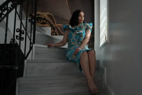 Elegant woman in a blue floral dress sitting on modern wooden stairs in a bright, moody interior.