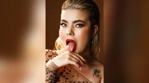 FiorellaRossi licks her lips with a wet tongue, showing her red lipstick and blonde hair against a brown background.