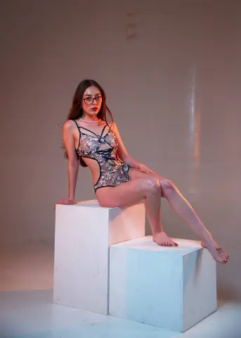 Young woman in a sheer black lace bodysuit sitting on a white pedestal in a minimalist studio setting.