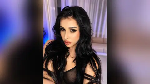 Glamorous woman with long wavy black hair wearing a black mesh top in a moody, dimly lit room.