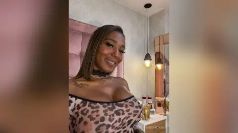 Smiling woman in a leopard print top and black lace choker posing in a glamorous pink bedroom setting.