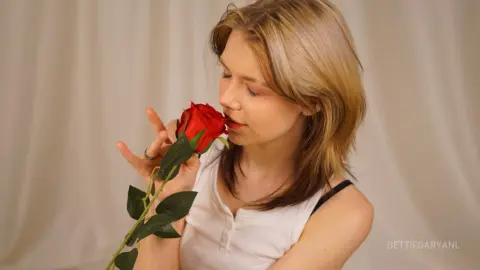 Blonde woman in a white tank top smelling a single red rose against a neutral background.