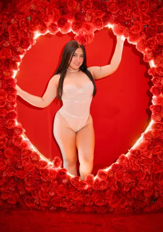 Smiling woman in white bodysuit posing inside a romantic red rose heart frame against a red background.