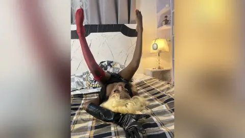 Blonde woman in black leather outfit and red stockings lying playfully on a plaid bed in a cozy bedroom.