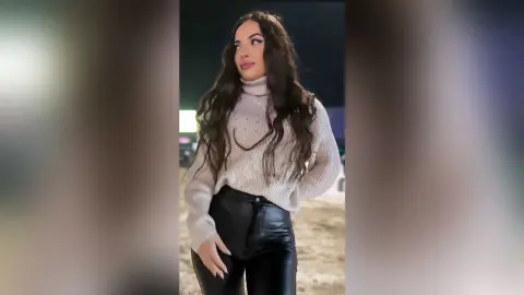 Smiling woman in a cozy beige turtleneck sweater and black leather pants posing outdoors at night.