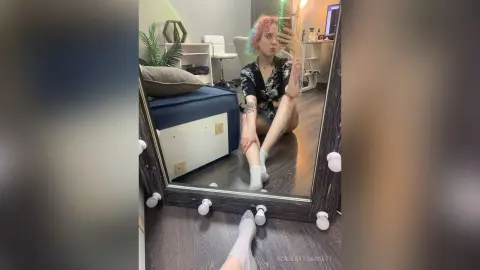 ScarlettGarrett sits on a dark blue chair in a black patterned top, looking into a mirror with her legs bent and white socks on.