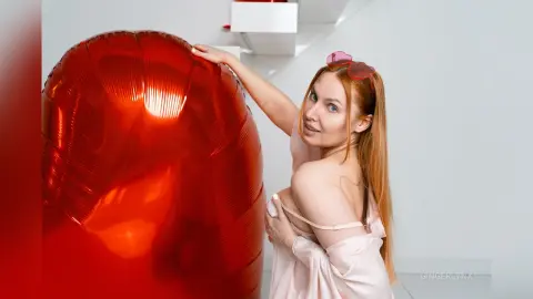 Smiling redhead woman in a pink silk top leaning against a large red balloon in a bright minimalist room.