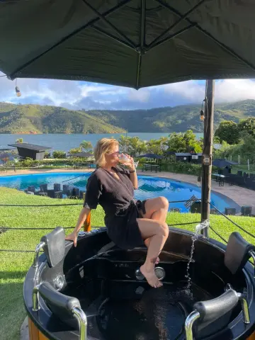 Relaxed woman in a black robe sipping a drink while sitting on the edge of a luxury outdoor hot tub by a lake.