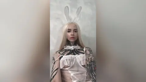 Blonde woman in a white lace corset and bunny ears posing in a dreamy, ethereal indoor setting.