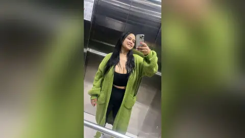 NiaCorelli poses in a mirror selfie wearing a long lime green cardigan over a black crop top and black leggings in an elevator.