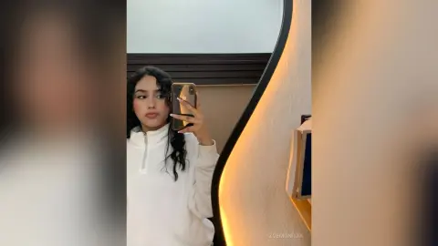 ZoeMonrow poses for a mirror selfie wearing a white long-sleeve zip-up top, showing off her long dark wavy hair.
