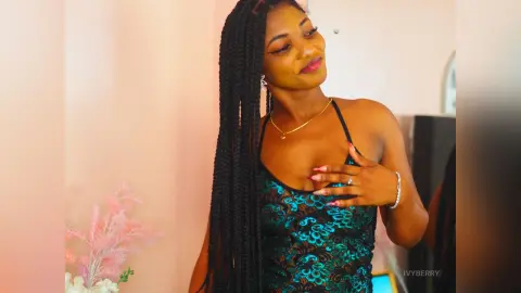 IvyBerry poses in a teal lace camisole, showing off her long black braids and gold jewelry against a soft pink background.