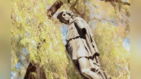 Classical stone statue of a young man in a draped tunic standing amidst lush green willow trees in soft sunlight.