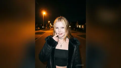 JaimieBez licks her finger while wearing a black crop top and a black furry jacket outdoors at night.
