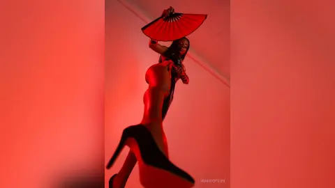 Stylish woman in a black bodysuit and high heels posing with a red fan under dramatic red lighting.